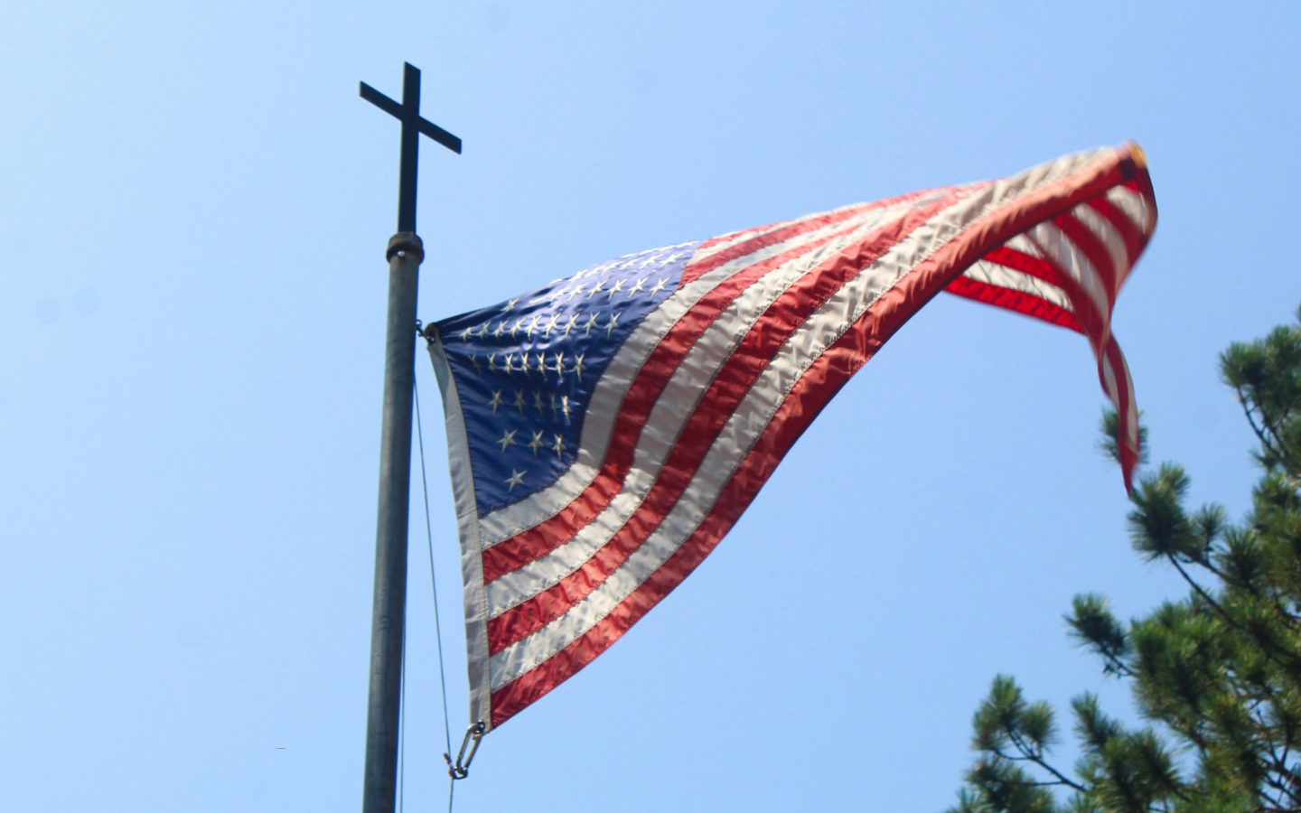 A U.S. flag waving from a flag pole with a cross on top.