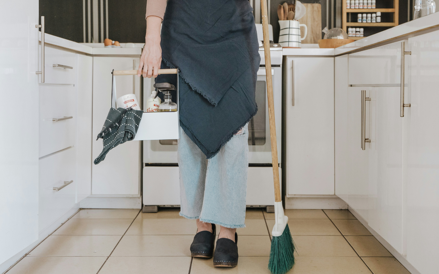 A lady carries cleaning materials into a kitchen.
