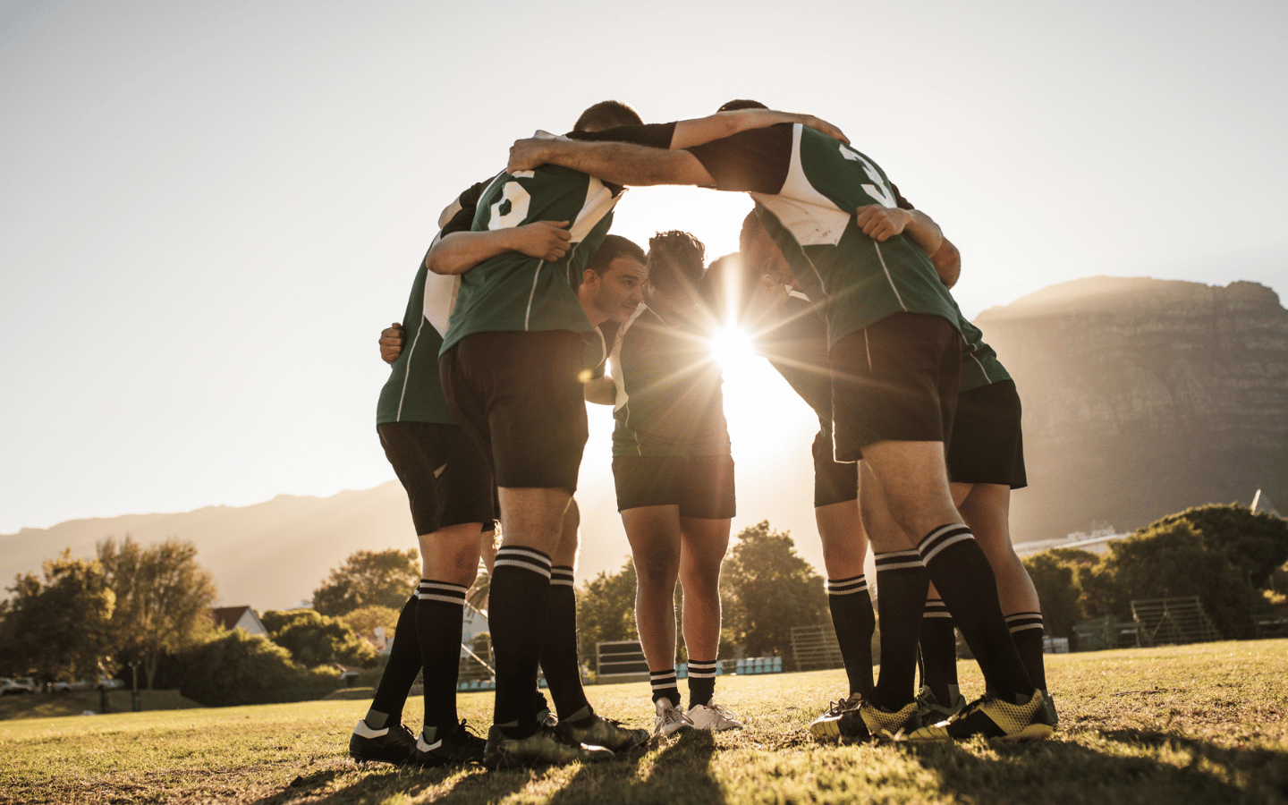 A rugby team gathers at midfield with the sun shining through their huddle.