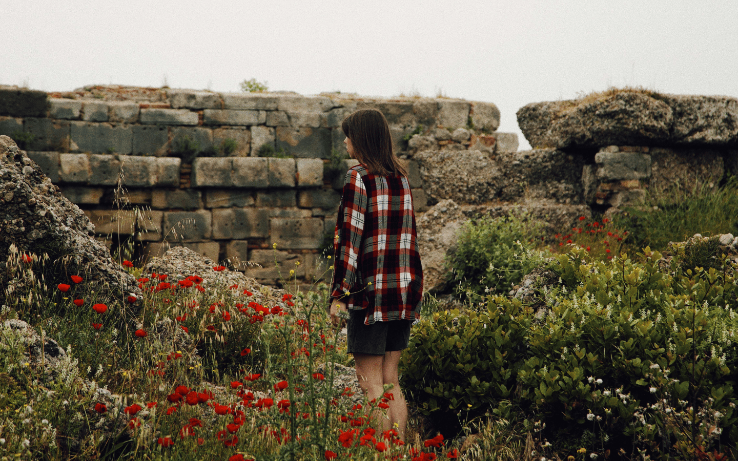 A woman walks through a patch of wildflowers growing among ruins.