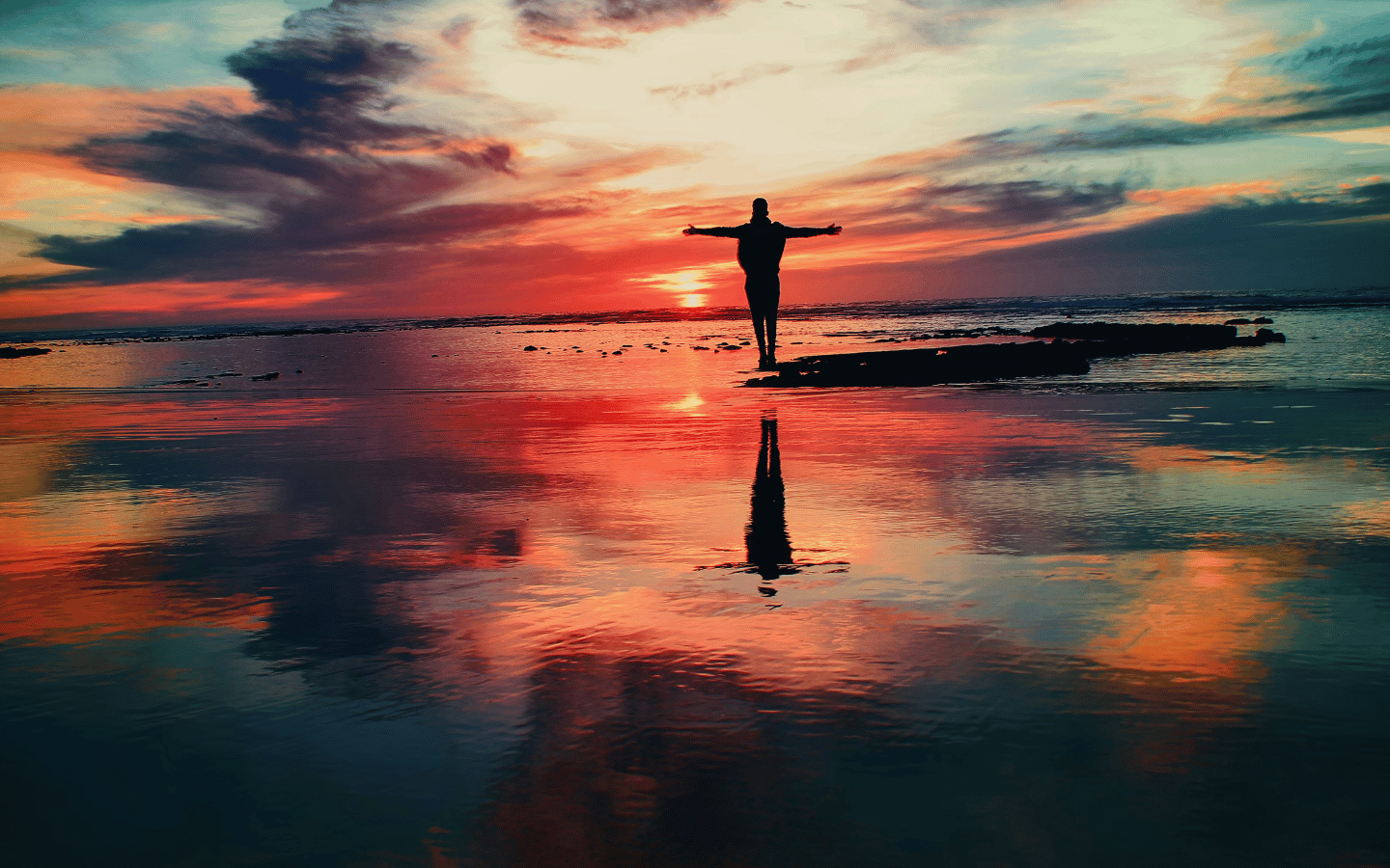 A woman stands on a beach during a colorful sunset with her arms stretched wide.