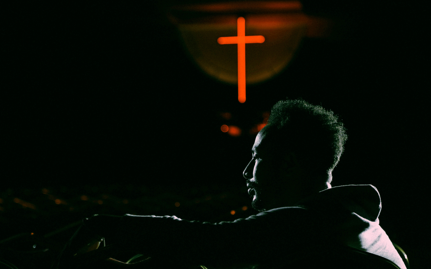 The profile of a man sitting under a neon cross, contemplating.