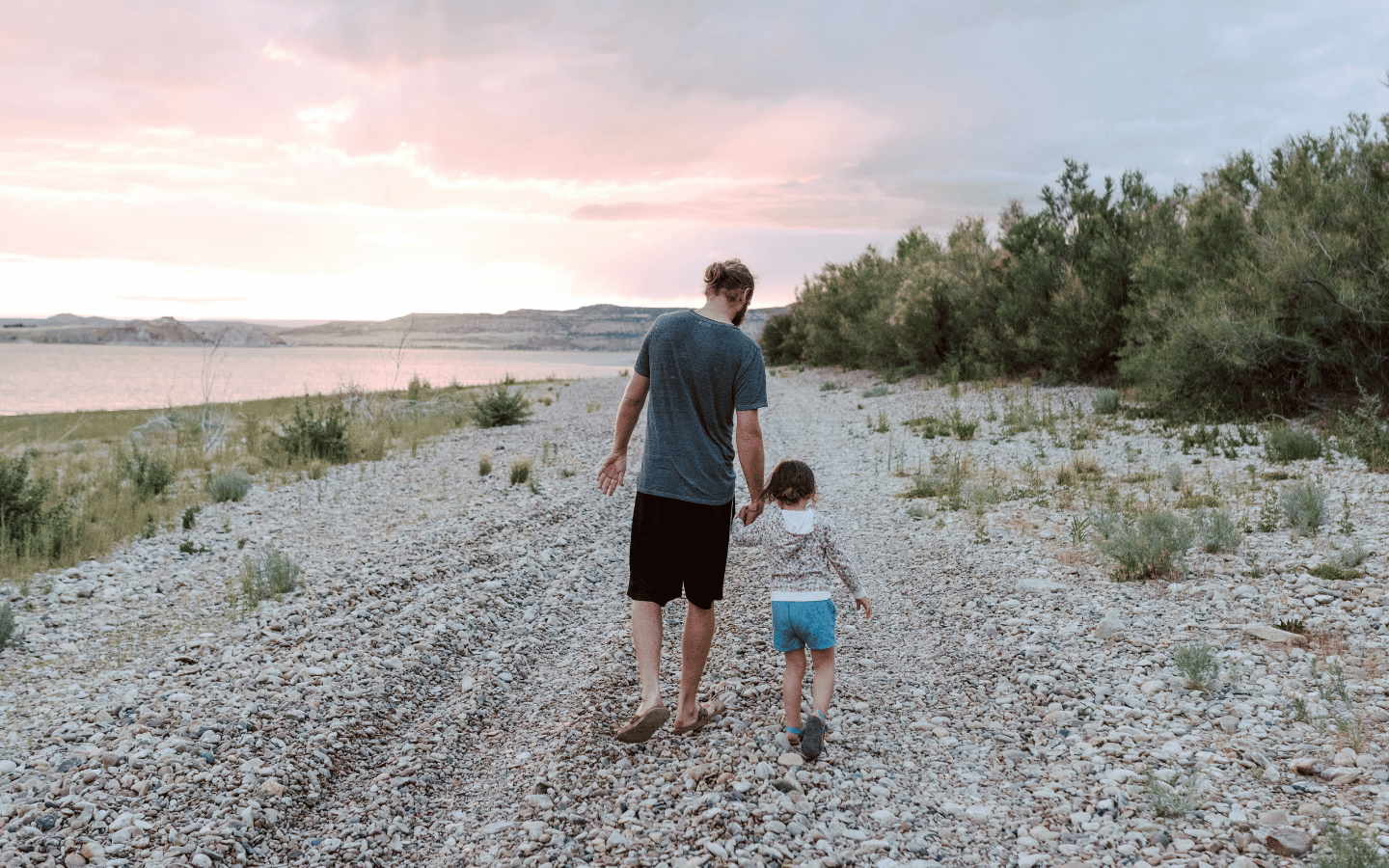 A man walks his young daughter along a gravel pathway near a body of water.