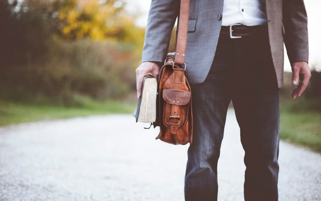 A cropped image of a man holding a Bible and a satchel while walking on a gravel road.