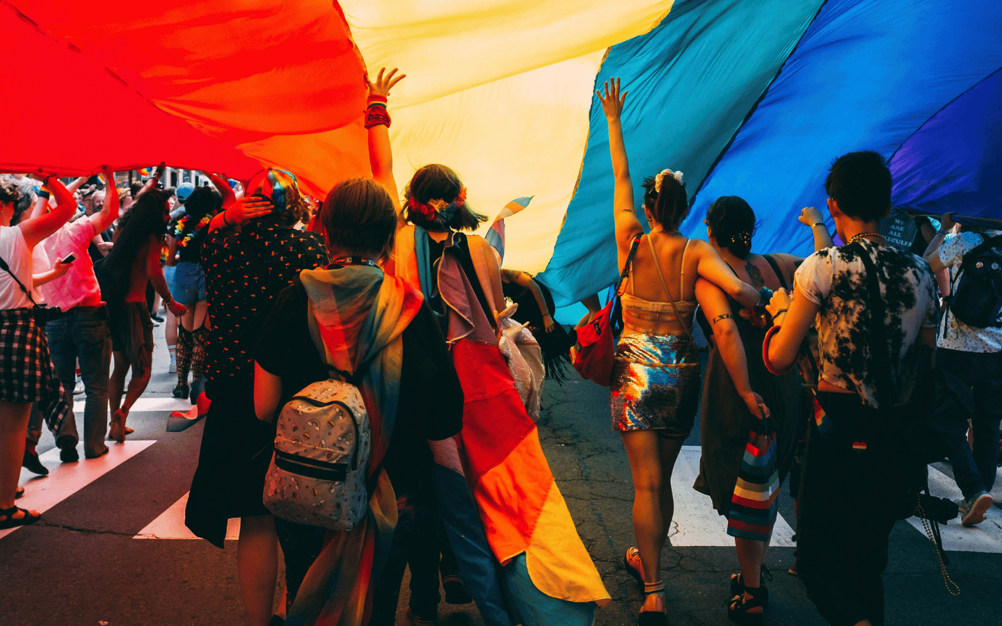 Young people walk in a Pride parade underneath a pride flag.