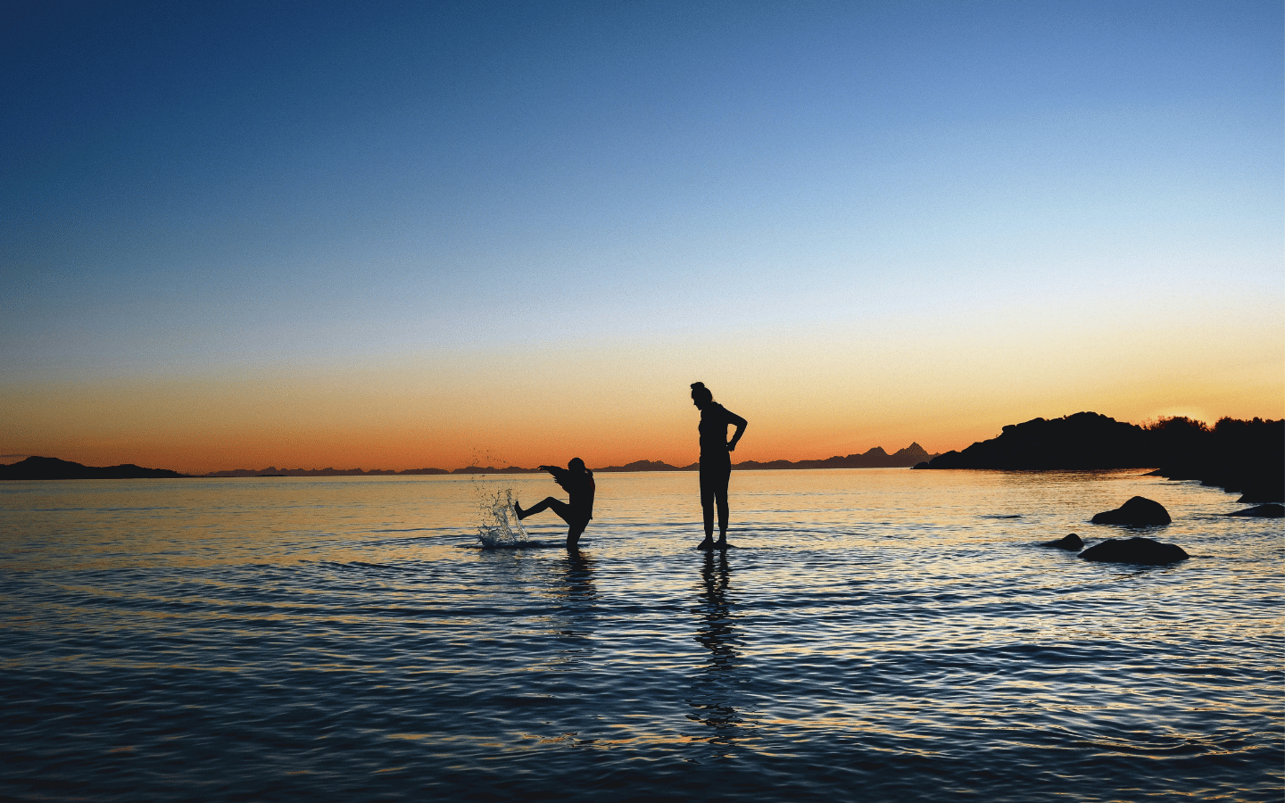 The silhouette of an adult watching a child splashing in an inlet of water they are standing in.