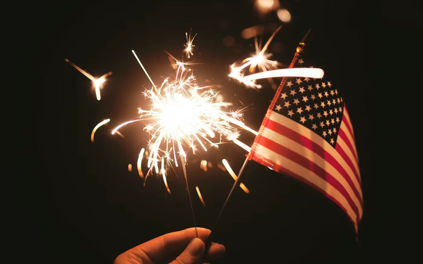A person’s hand holding a sparkler and a flag at night.