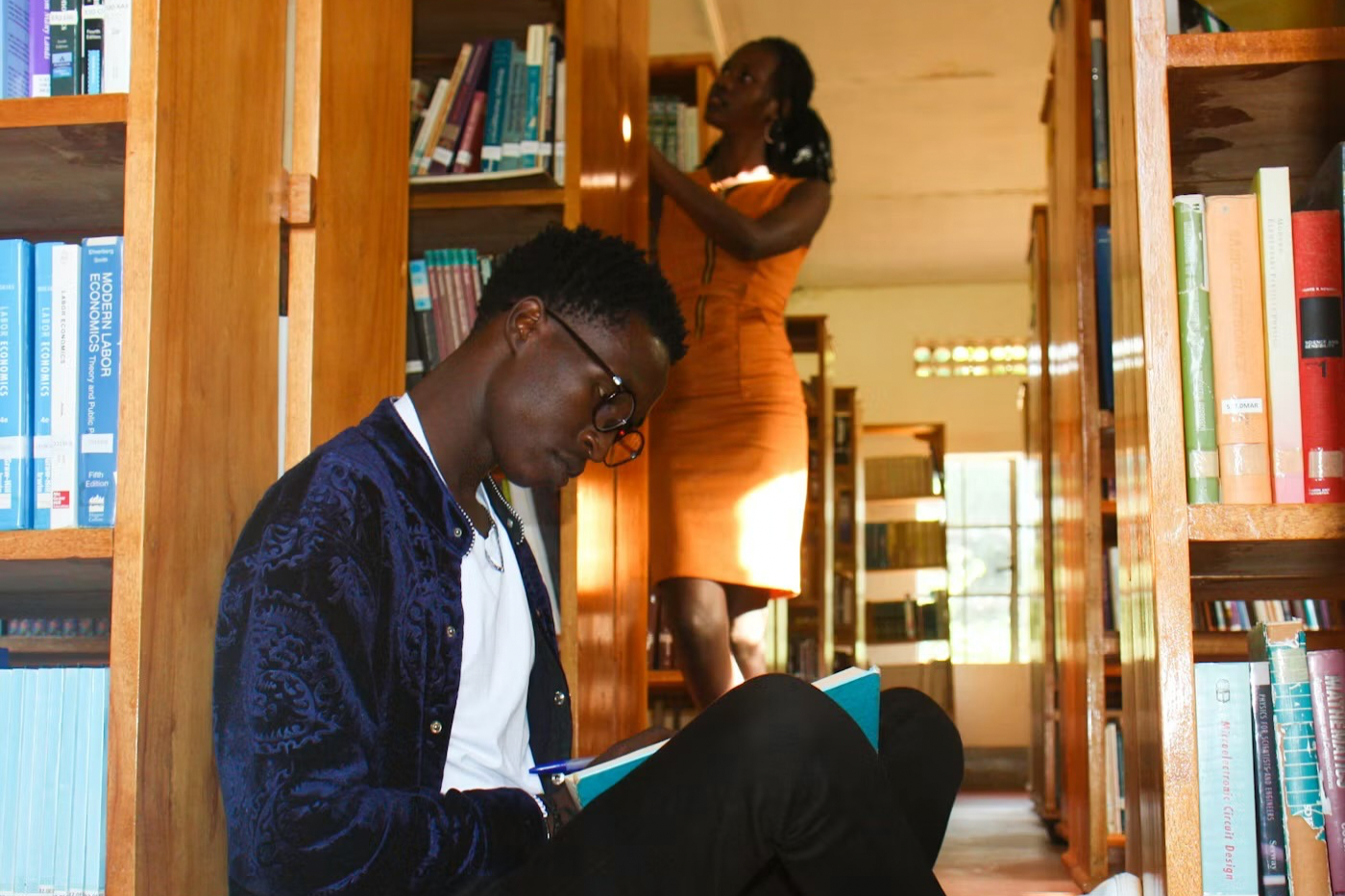 Two students in the stacks of a library, one is reading and another is searching for a book title.